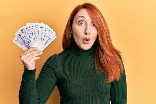 Beautiful redhead woman holding 2 singapore dollars banknotes scared and amazed with open mouth for surprise, disbelief face
