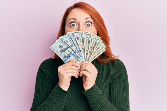 Beautiful redhead woman holding dollars close to face afraid and shocked with surprise and amazed expression, fear and excited face.