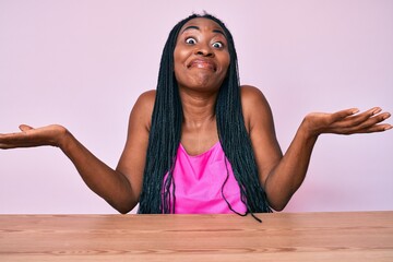 African american woman with braids wearing casual clothes sitting on the table clueless and confused expression with arms and hands raised. doubt concept.