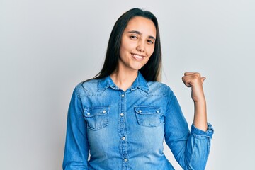Young hispanic woman wearing casual denim jacket smiling with happy face looking and pointing to the side with thumb up.