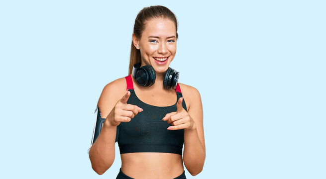Beautiful blonde woman wearing gym clothes and using headphones pointing fingers to camera with happy and funny face. good energy and vibes.