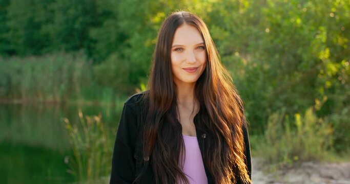Portrait of a smiling young woman with long dark hair outdoors in a forest near a lake.