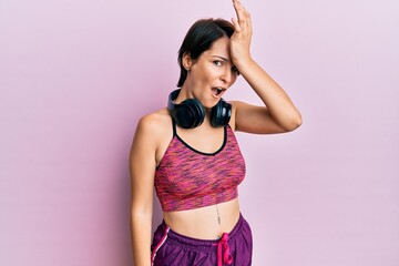 Young brunette woman with short hair wearing sportswear and headphones surprised with hand on head...