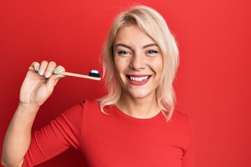 Young blonde girl holding toothbrush with toothpaste looking positive and happy standing and smiling with a confident smile showing teeth