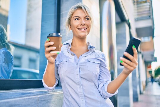 Young blonde woman smiling happy using smartphone and drinking take away coffee at the city.