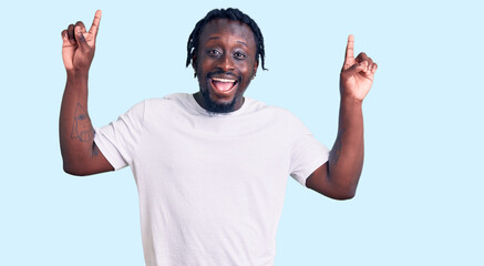 Fototapeta premium Young african american man with braids wearing casual white tshirt smiling amazed and surprised and pointing up with fingers and raised arms.