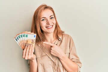 Young caucasian woman holding bunch of 50 euro banknotes smiling happy pointing with hand and finger