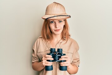 Young caucasian woman wearing explorer hat looking through binoculars skeptic and nervous, frowning...