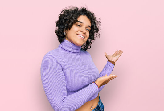 Young hispanic woman with curly hair wearing casual clothes inviting to enter smiling natural with open hand