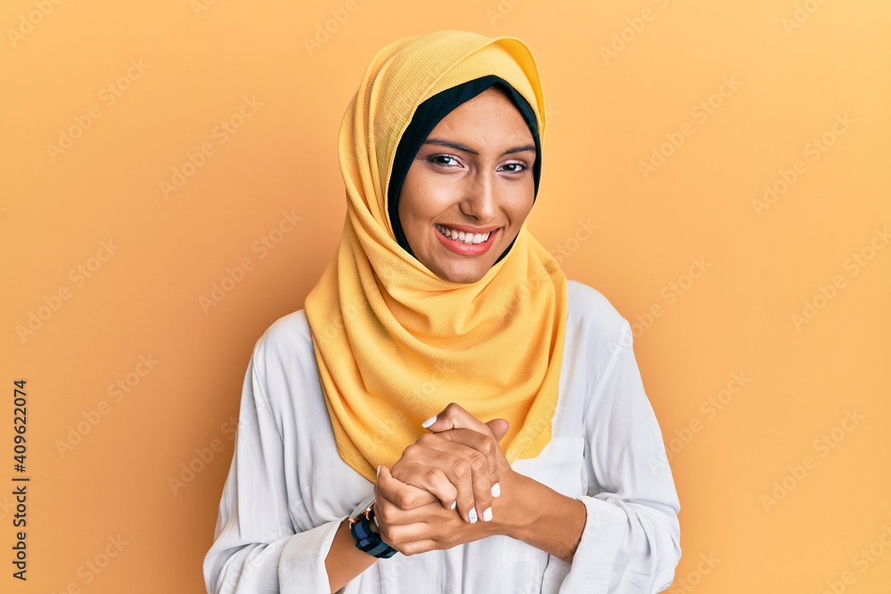 Poster Young brunette arab woman wearing traditional islamic hijab scarf with hands together and crossed fingers smiling relaxed and cheerful. success and optimistic