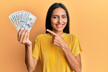 Young brunette woman holding dollars smiling happy pointing with hand and finger