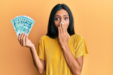 Young brunette woman holding 50 yuan chinese banknotes covering mouth with hand, shocked and afraid...