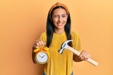 Young brunette woman holding alarm clock and hammer winking looking at the camera with sexy expression, cheerful and happy face.