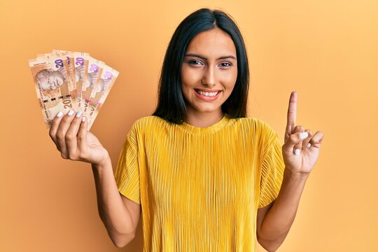 Young brunette woman holding south african 20 rand banknotes smiling with an idea or question pointing finger with happy face, number one