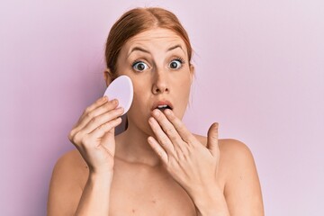 Young irish woman holding makeup sponge covering mouth with hand, shocked and afraid for mistake. surprised expression