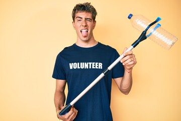 Young handsome man recycling plastic bottle holding litter pick up tool sticking tongue out happy with funny expression.