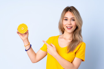 Teenager Russian girl holding an orange isolated on blue background and pointing it