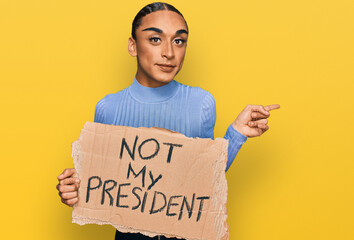 Hispanic transgender man wearing make up and long hair holding not my president protest banner smiling happy pointing with hand and finger to the side