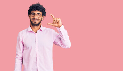 Handsome young man with curly hair and bear wearing business clothes smiling and confident gesturing with hand doing small size sign with fingers looking and the camera. measure concept.