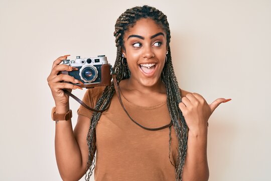 Young african american woman with braids holding vintage camera pointing thumb up to the side smiling happy with open mouth