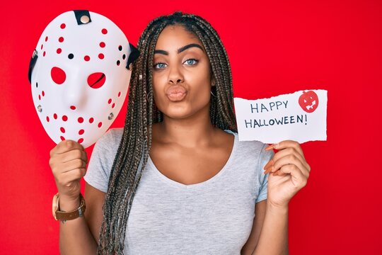 Young african american woman with braids holding halloween banner and scary mask looking at the camera blowing a kiss being lovely and sexy. love expression.