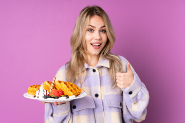 Teenager Russian girl holding waffles isolated on purple background showing ok sign and thumb up gesture