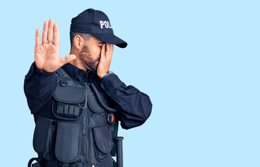 Young hispanic man wearing police uniform covering eyes with hands and doing stop gesture with sad...