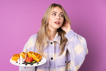 Teenager Russian girl holding waffles isolated on purple background listening to something by putting hand on the ear