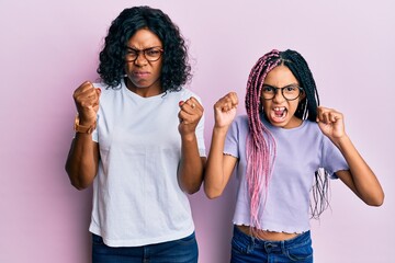 Fototapeta na wymiar Beautiful african american mother and daughter wearing casual clothes and glasses angry and mad raising fists frustrated and furious while shouting with anger. rage and aggressive concept.