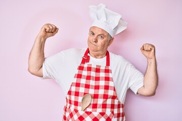 Senior grey-haired man wearing professional baker apron showing arms muscles smiling proud. fitness concept.