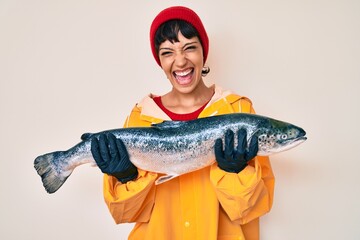 Beautiful brunettte fisher woman wearing raincoat holding fresh salmon smiling and laughing hard...