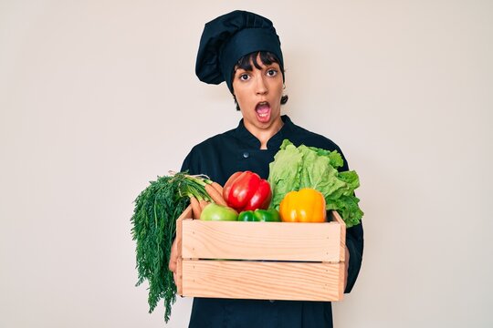 Beautiful brunettte woman chef holding fresh veggetables in shock face, looking skeptical and sarcastic, surprised with open mouth