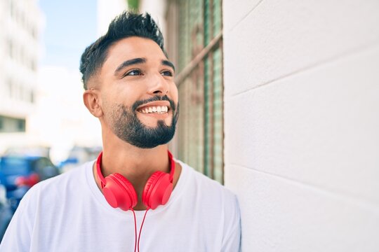 Young arab man smiling happy using headphones leaning on the wall at the city.