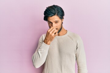 Young hispanic man wearing casual winter sweater smelling something stinky and disgusting,...