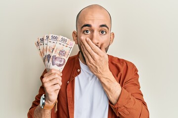 Young hispanic man holding 500 mexican pesos banknotes covering mouth with hand, shocked and afraid...