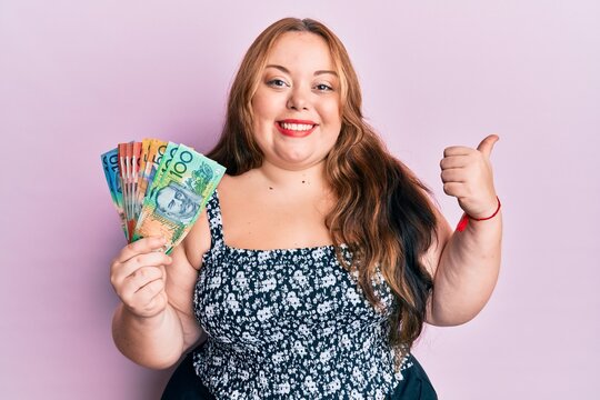 Plus size caucasian young woman holding australian dollars pointing thumb up to the side smiling happy with open mouth