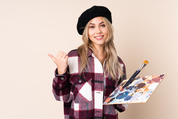 Teenager artist girl holding a palette isolated on blue background pointing to the side to present a product