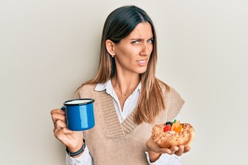 Brunette young woman drinking coffee and eating pastry clueless and confused expression. doubt concept.