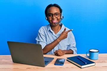 Young african woman wearing call center agent headset smiling cheerful pointing with hand and finger up to the side
