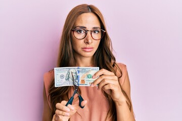 Young brunette woman cutting dollars with scissors for currency devaluation relaxed with serious...