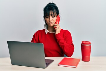 Young brunette woman with bangs working at the office talking on the smartphone depressed and worry...