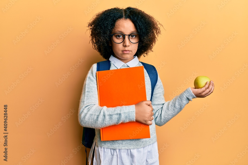 Sticker Young little girl with afro hair wearing school bag holding books and green apple relaxed with serious expression on face. simple and natural looking at the camera.