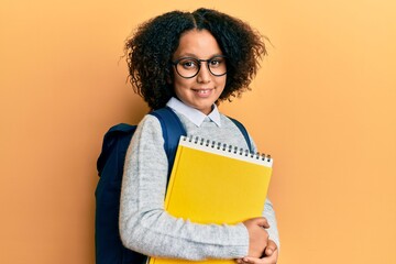 Young little girl with afro hair wearing school bag and holding books looking positive and happy standing and smiling with a confident smile showing teeth