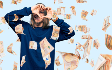 Handsome caucasian man with long hair wearing casual winter sweater doing heart shape with hand and fingers smiling looking through sign