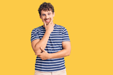 Young handsome man with curly hair wearing casual clothes looking confident at the camera smiling with crossed arms and hand raised on chin. thinking positive.