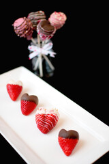Heart Shape Strawberry Covered Chocolate decorated with sprinkles on white rectangle plate with strawberry pop on black background