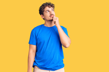 Young handsome man with curly hair wearing casual clothes with hand on chin thinking about...