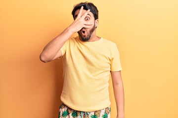 Young hispanic man wearing swimwear peeking in shock covering face and eyes with hand, looking...