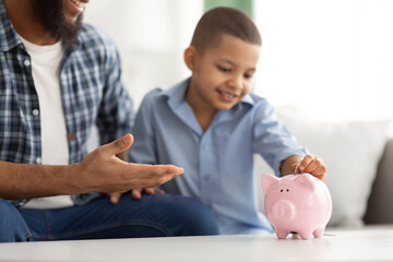 Black Dad And Son Putting Coin In Piggybank Indoors, Cropped