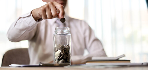 A man with a coin is about to drop it into a glass bottle with a lot of coins inside, he saves...